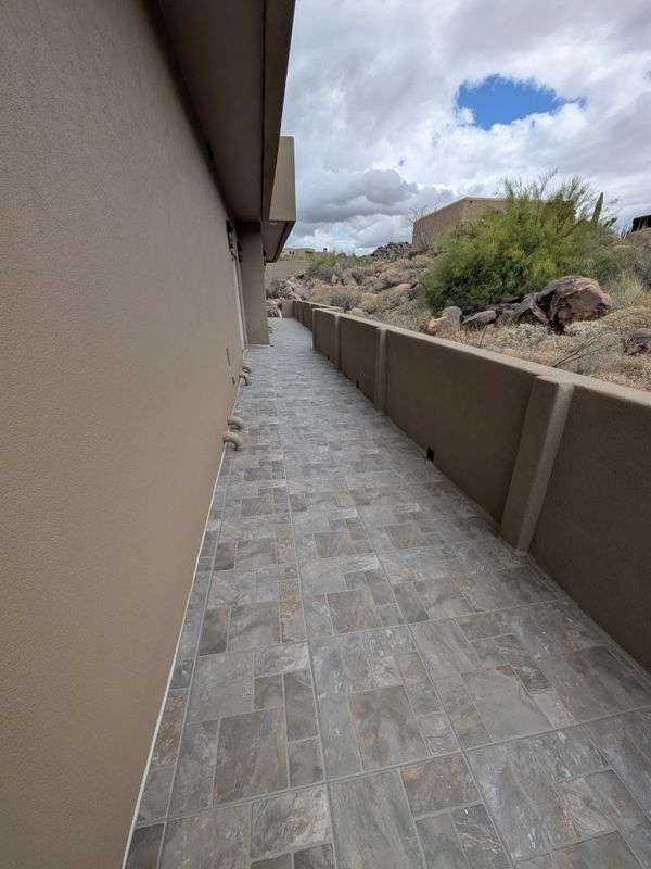 Narrow tiled walkway alongside a beige building with a low wall and desert landscape.