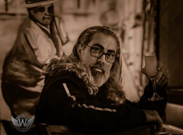 Man with long hair and glasses holding a glass of champagne.