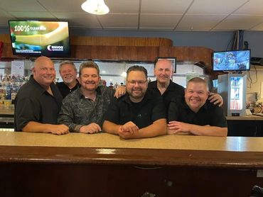 Group of six men smiling behind a bar counter.