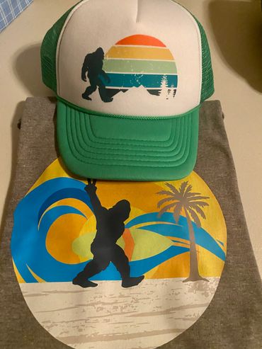 Green cap and gray shirt with Bigfoot graphics in sunset and beach themes.