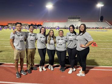 Group of seven people wearing matching 'Boo Boo Crew' shirts at a sports field during sunset.