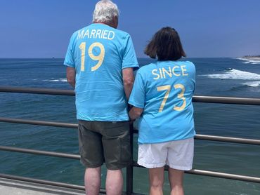 Elderly couple wearing matching shirts by the ocean, celebrating marriage since 1973.