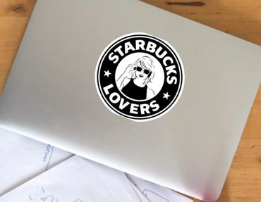 Laptop with a 'Starbucks Lovers' sticker on a wooden desk surrounded by sketches and pens.