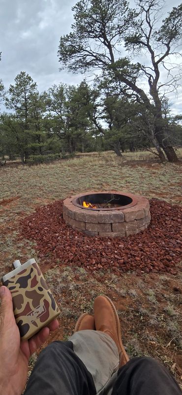 Person relaxing by a small fire pit in a forest holding a camo flask.