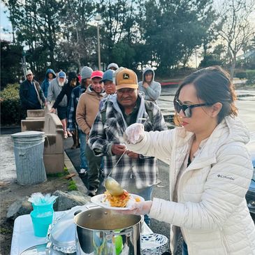 A woman serves hot food to people waiting in line outdoors.