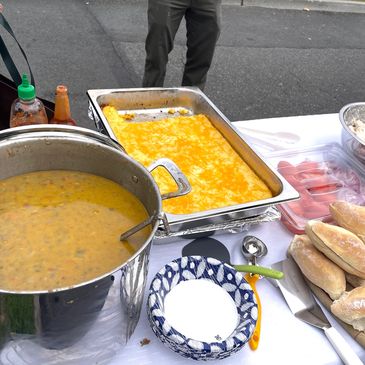 Outdoor food setup with soup, baked dish, and bread rolls.