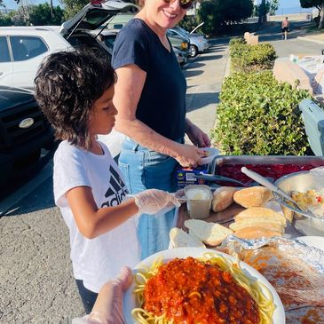 Child and woman serving spaghetti at outdoor event.