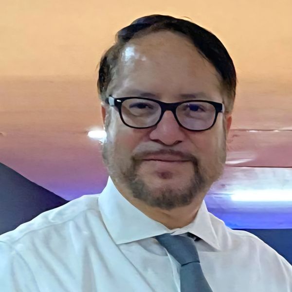 Man in glasses, white shirt, and gray tie smiling indoors.