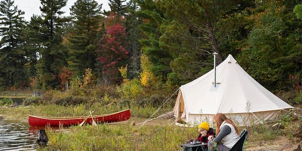Two people playing chess near a tent by a lakeside with a red canoe and forest in the background.