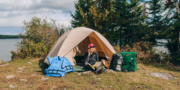 A woman sitting at a lakeside campsite with a tent and outdoor gear.