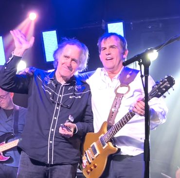 Two musicians on stage, one waving and the other holding a guitar.