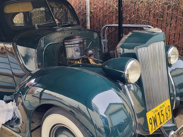 A vintage dark green car with a 1938 California license plate and its hood open.