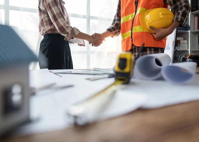 Two professionals shaking hands over architectural plans and construction tools on a desk.