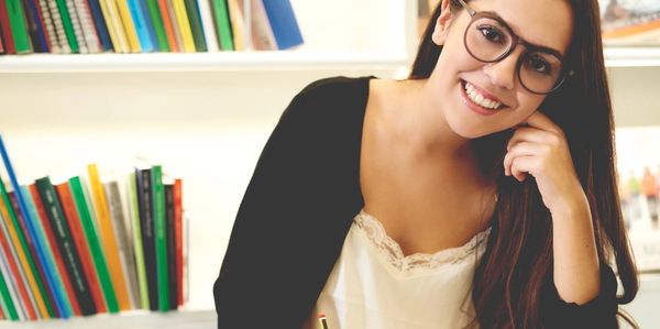 Smiling attractive young student wearing glasses sitting cross legged studying on the floor 