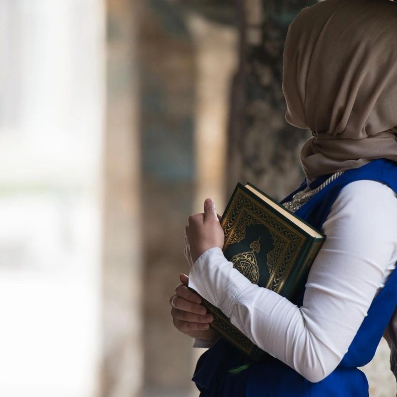 Woman in hijab holding a Quran, standing thoughtfully.