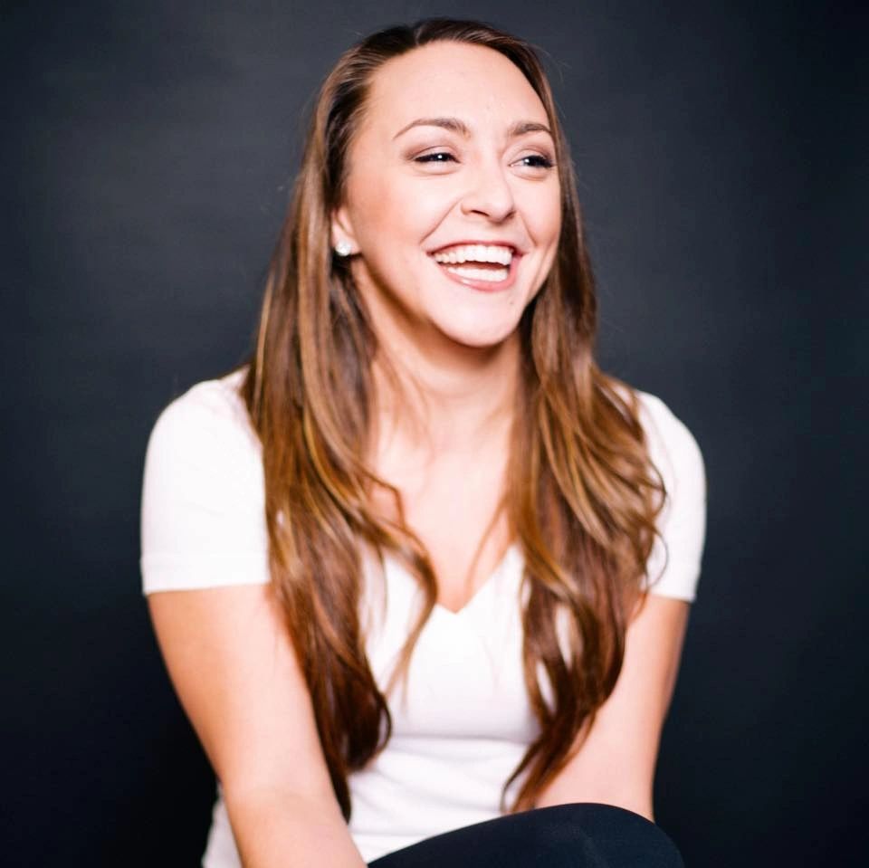 A woman with long, dark hair sits smiling big, as if she were laughing.