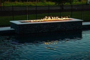 Modern rectangular fire pit beside a pool with flames reflecting in the water.