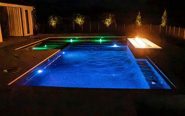 A modern pool illuminated with blue and green lights and a fire pit at night.