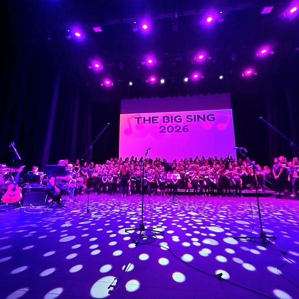A large choir on stage for The Big Sing 2026 event under purple lights.