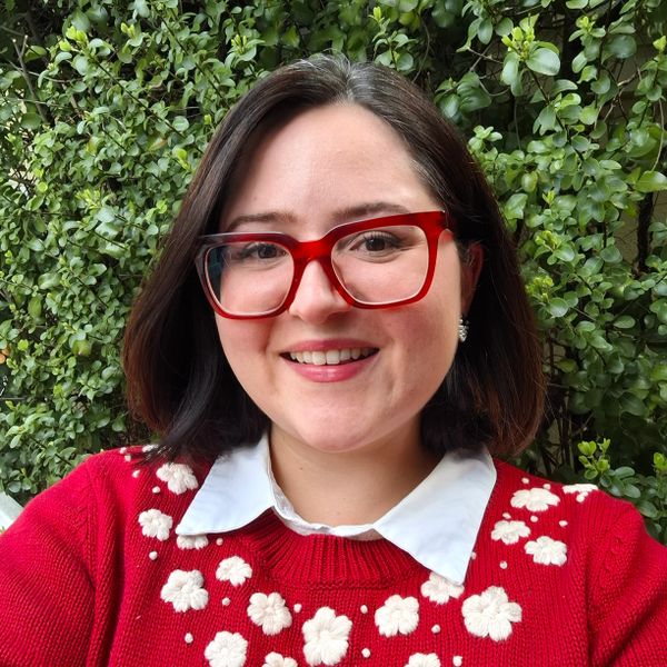 Smiling woman with red glasses and floral sweater in front of green foliage.