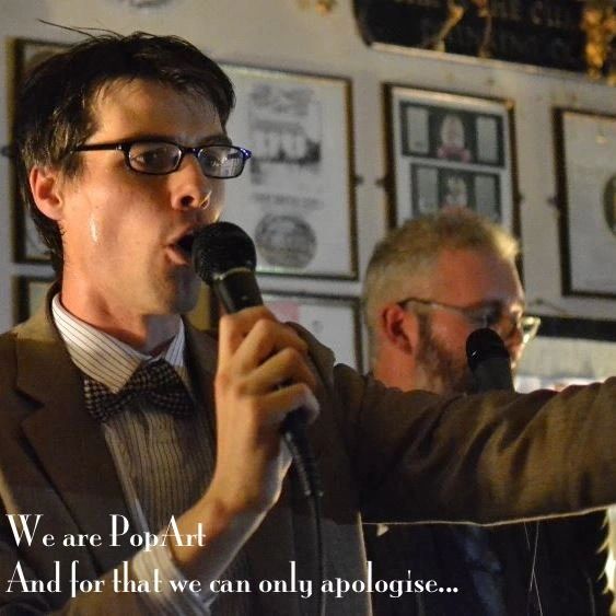 Two men speaking into microphones, one wearing glasses and a bow tie.