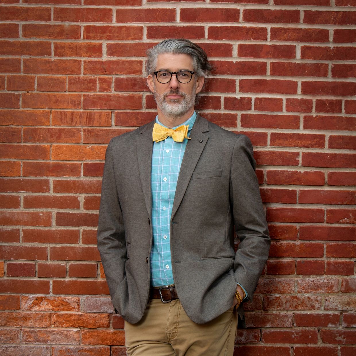 Man in a gray blazer and yellow bow tie stands against a red brick wall.