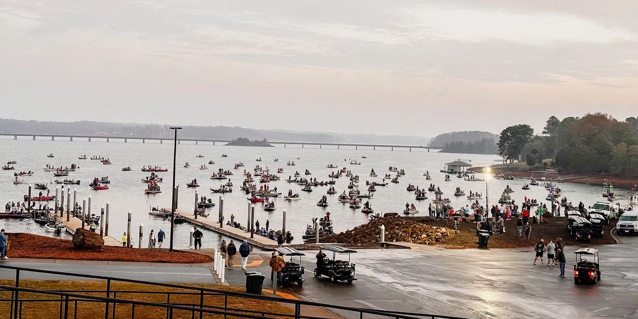 Many boats are gathered on a lake near docks with people onshore.