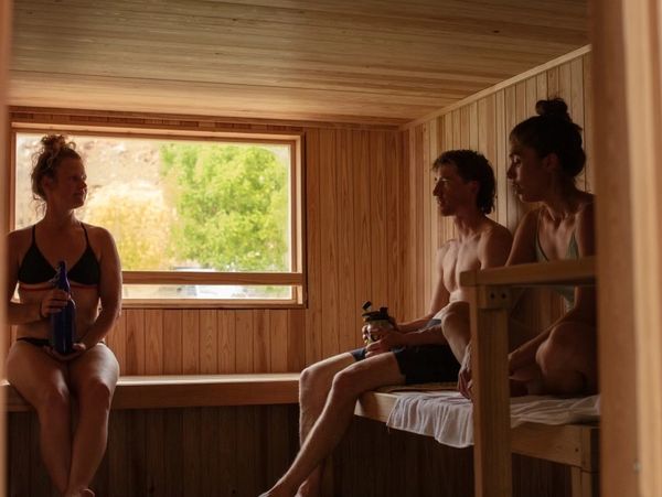 Three people relaxing in a wooden sauna with a window showing greenery outside.