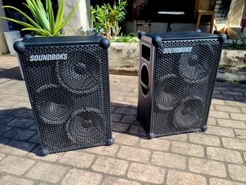 Two black SOUNDBOKS speakers on a stone patio.