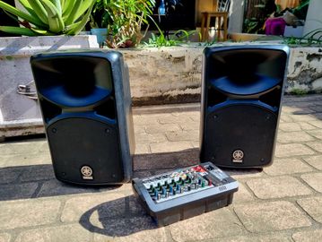 Two black speakers and a mixer on an outdoor surface.
