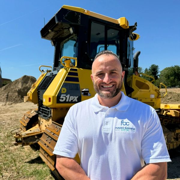 Man in white polo shirt stands smiling in front of a yellow bulldozer on a sunny day.