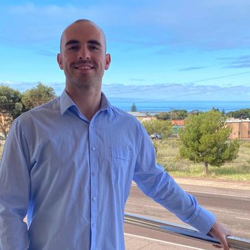 Smiling man in a blue shirt standing outdoors with a scenic background.