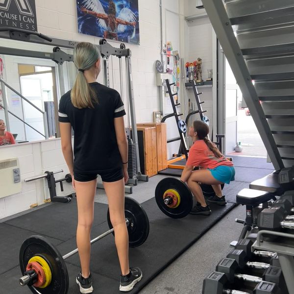 Two girls lifting barbells in a gym, one squatting and the other standing.