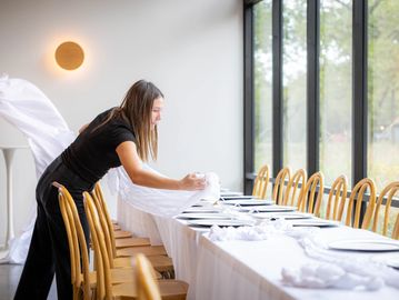 Woman setting a table in a bright dining room with wooden chairs.