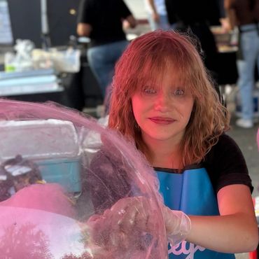 Two women making cotton candy at a colorful market stall with various flavored toppings.