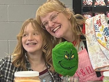 Two women smiling behind a colorful cotton candy stand with various flavors displayed.