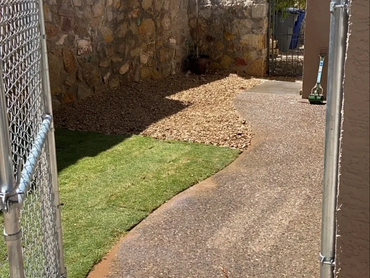 A backyard with stone walls, gravel, grass, and a metal gate under a clear sky.