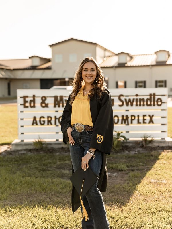 Young woman in a graduation gown holding a cap outdoors at an agricultural complex.
