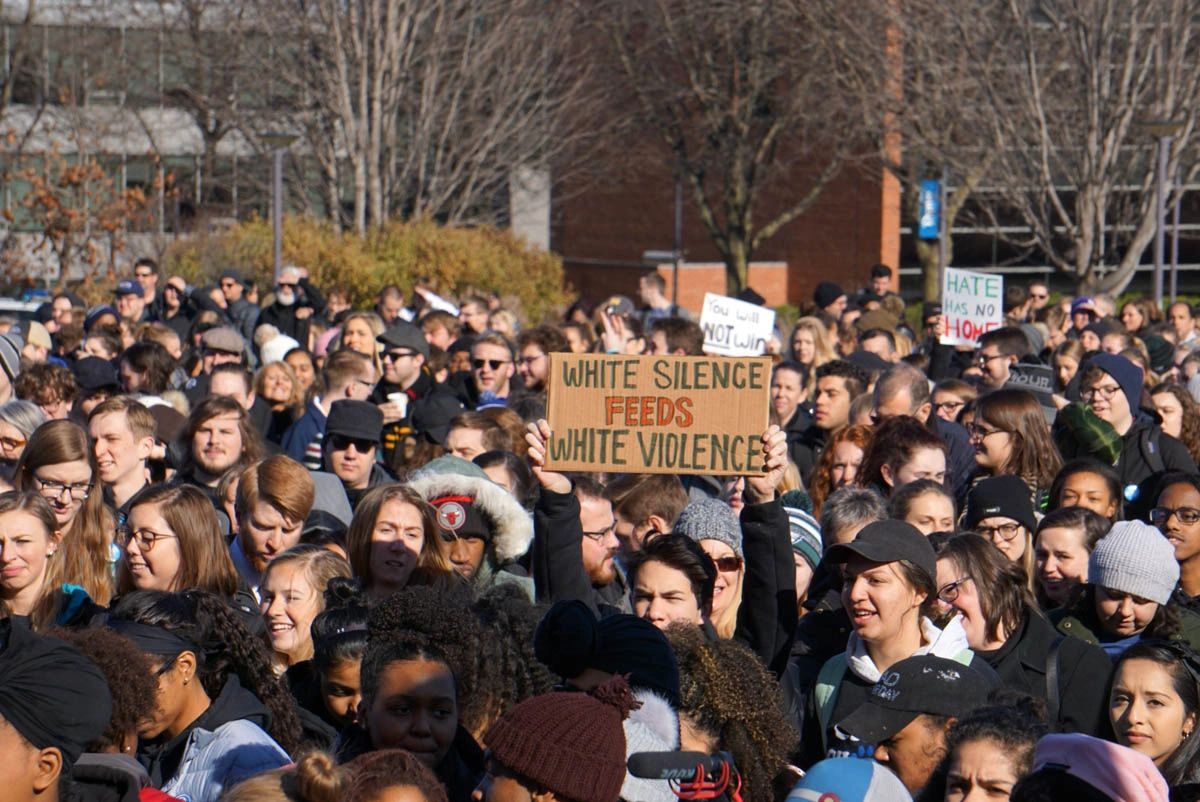 Rally crowd with one person holding up a cardboard sign saying White Silence Feeds White Violence