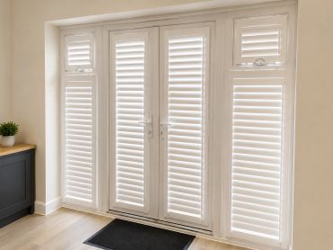 White shutter blinds covering glass doors and windows in a bright room.