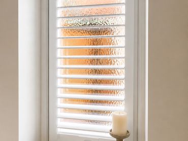 White plantation shutters covering a textured glass window with a candle on the sill.