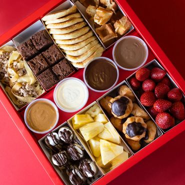 A box with different chocolate dips, brownies, pancakes, waffles, pineapple, and strawberries
