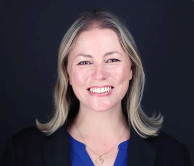 Image of a smiling blonde woman with a blue shirt, black jacket and black background.