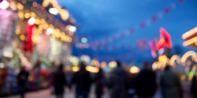 Blurred image of a crowded outdoor fair or festival at dusk with colorful lights.