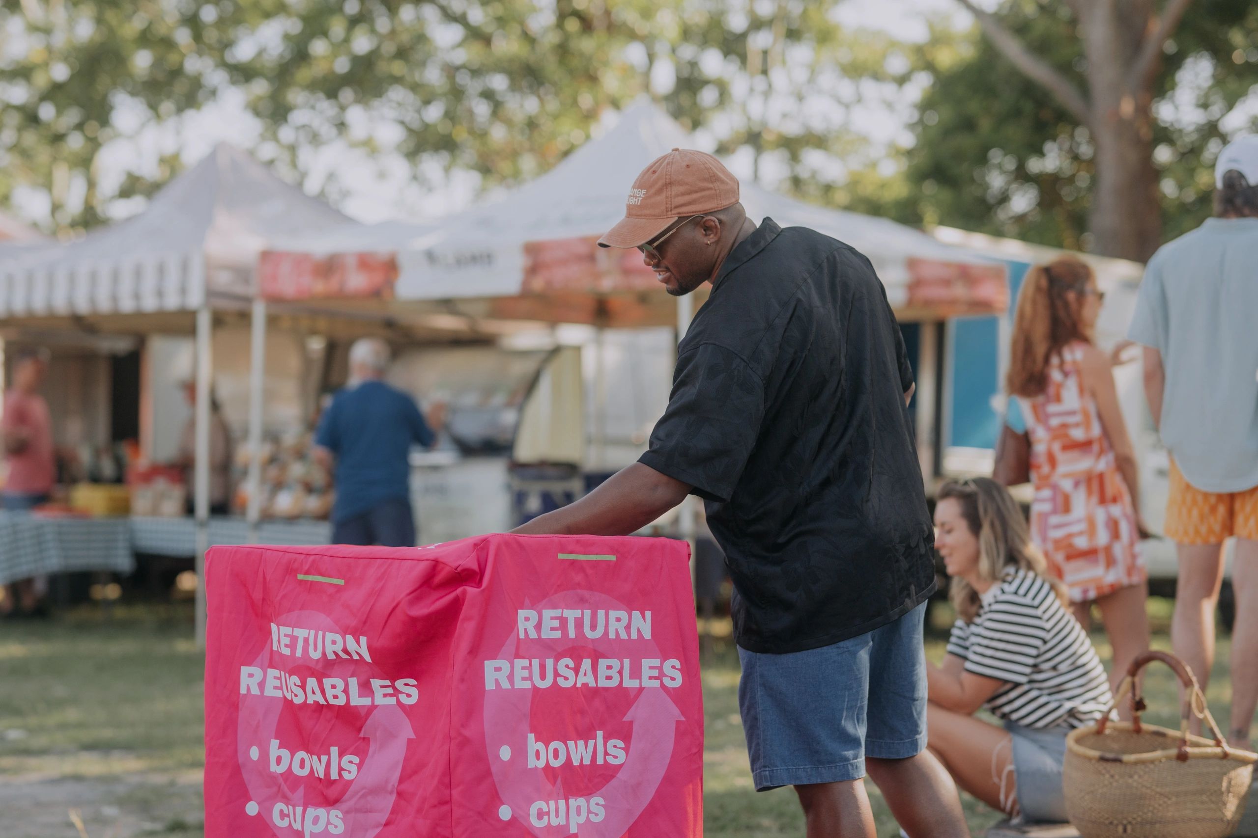 Man wearing a cap at an outdoor event near reusable item bins.
