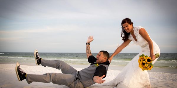Bride playfully drags groom on a sandy beach during wedding photos.