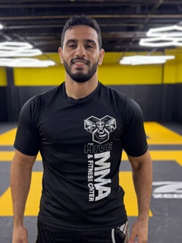 A man in MMA gym attire standing on a yellow and gray mat, smiling.