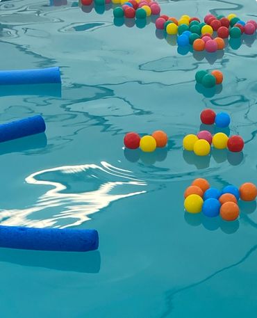 Colorful balls and blue pool noodles float on water's surface.