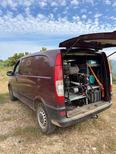 A maroon van with open back filled with equipment in a rural setting.