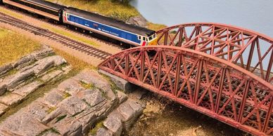 Model train crossing a red bridge on a detailed miniature landscape.